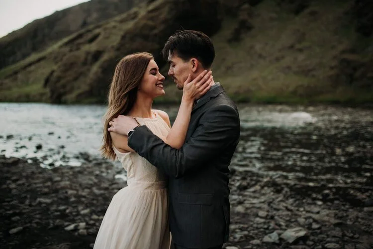A young woman in a white dress and a man in a dark suit holding each other close by a river with a mountainous landscape in the background.