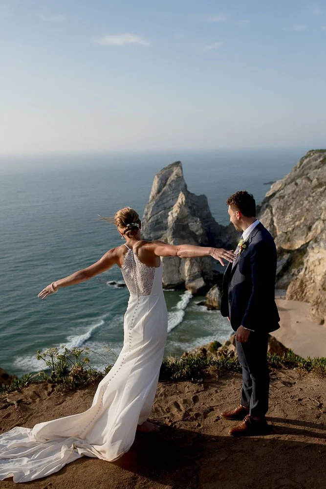 Bride in a white wedding dress extending her arms towards groom in a dark suit on a cliff overlooking the ocean.