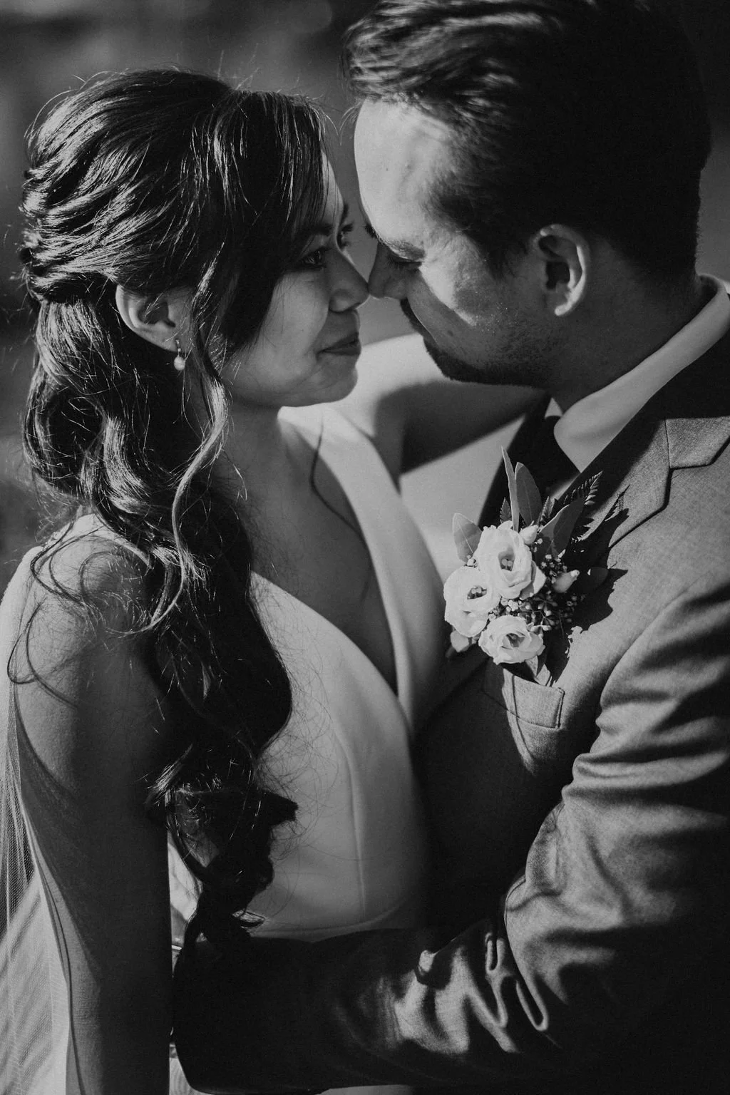A black-and-white photo of a couple on their wedding day, facing each other closely with noses touching, smiling softly. The woman has long, wavy hair and is wearing earrings and a dress, while the man is dressed in a suit with a boutonniere featurin