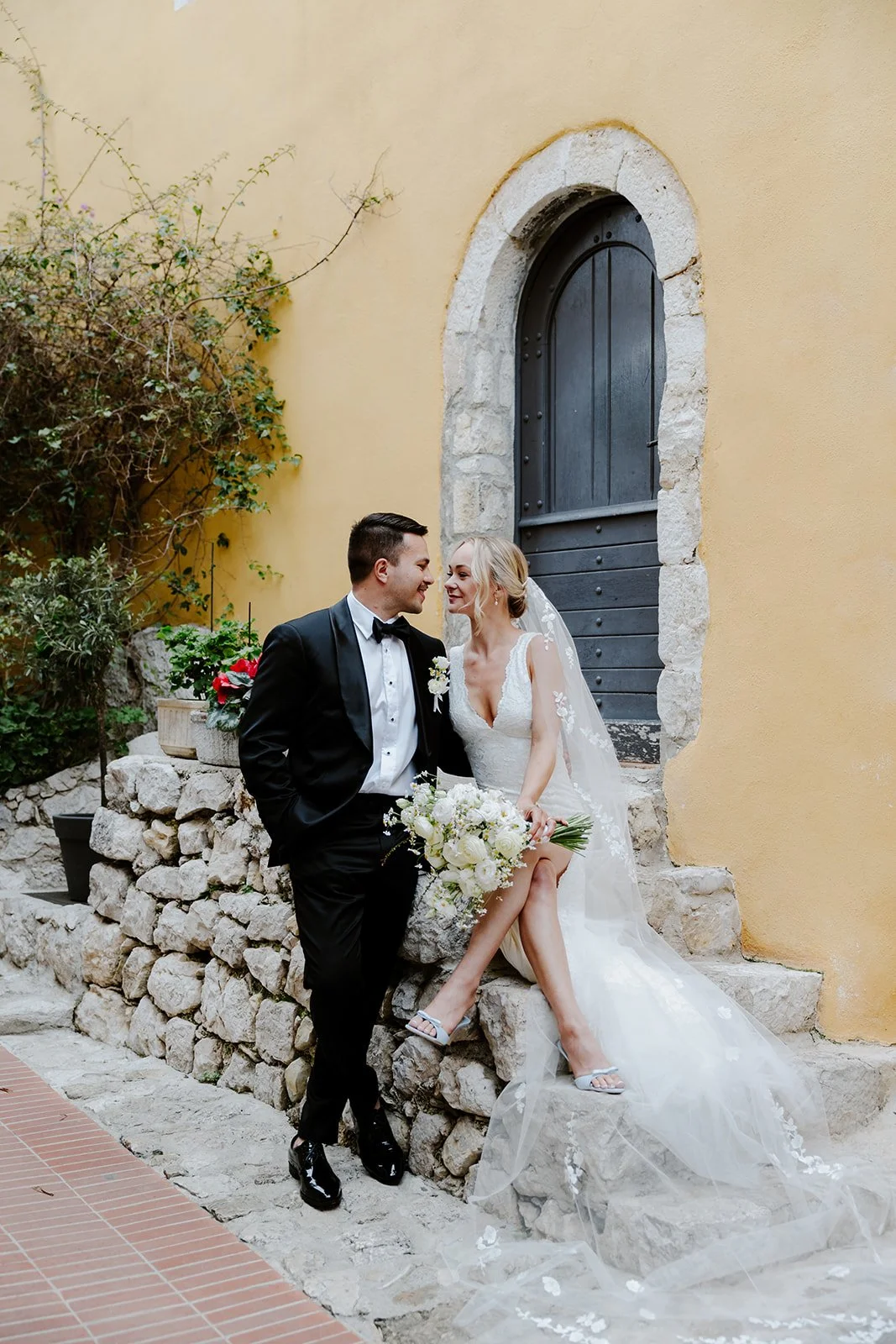A bride and groom on their wedding day, sitting on a stone wall outside a yellow building with a black arched door, sharing an affectionate moment with the bride holding a large bouquet of white flowers.