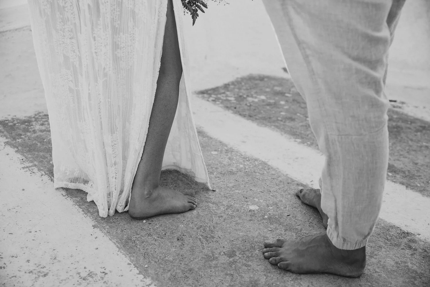 Black and white photo of a couple standing barefoot on sand, with the woman wearing a long, flowing dress and the man in light-colored pants. Their feet are close together, and only the lower part of their bodies is visible.