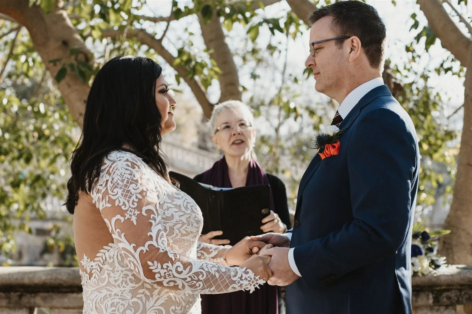 A couple wedding ceremony outdoors, holding hands and facing each other, with a woman officiant standing behind them in front of trees.