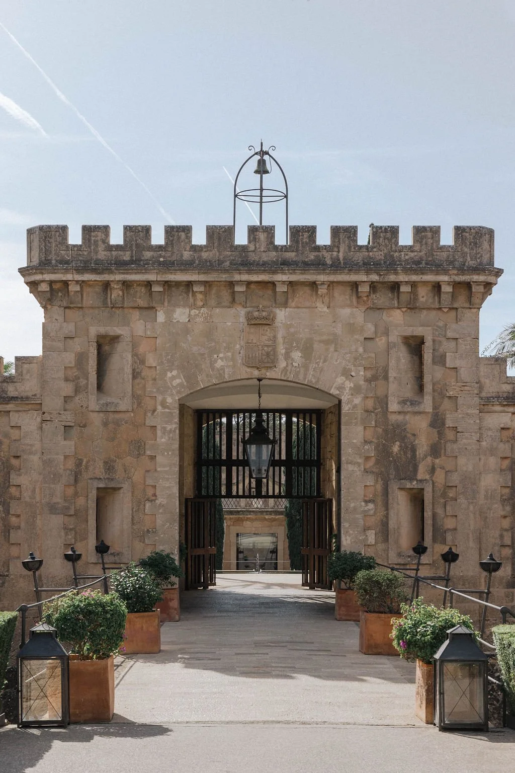 Historic stone gate with wooden doors, potted plants, lanterns, and a guard tower with a bell on top.
