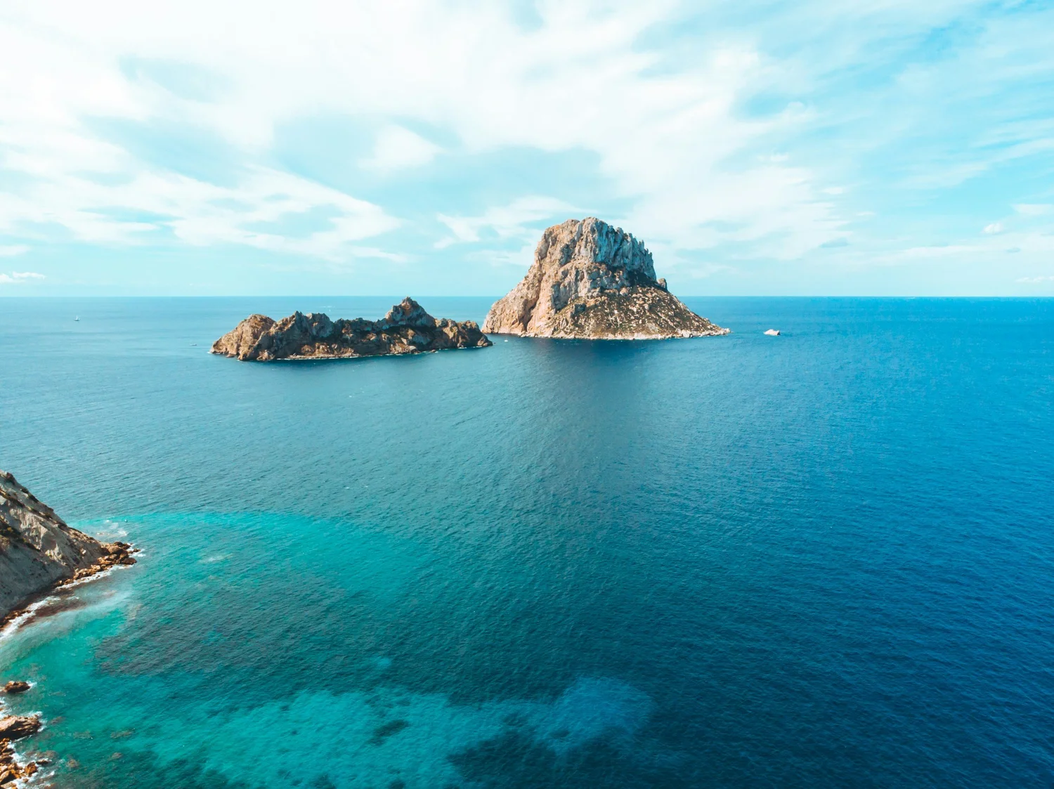A large rock formation in the middle of the ocean with smaller rocks nearby, under a partly cloudy sky. The water is a vibrant blue with areas of turquoise near the shore.