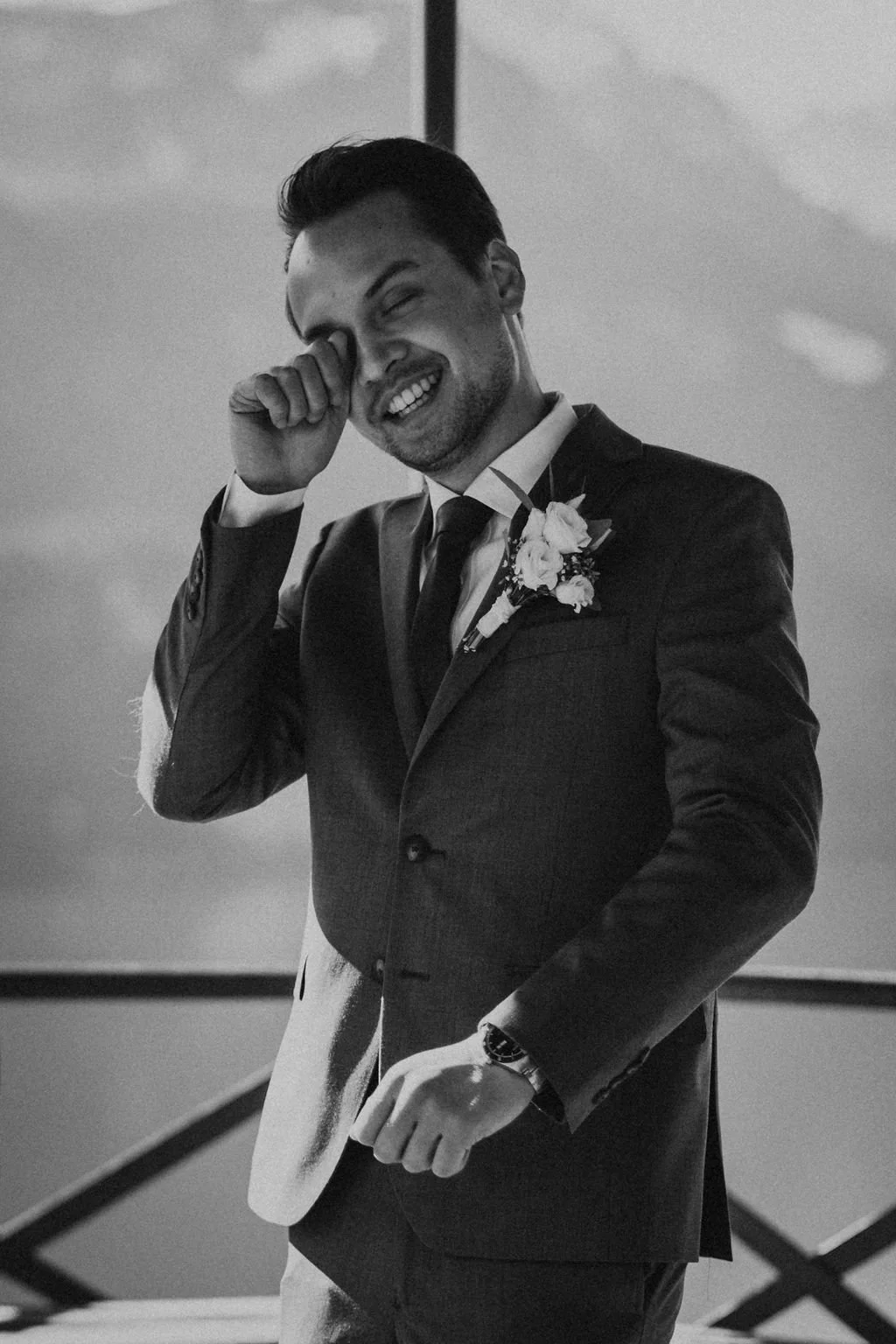 A groom in a suit with a boutonniere, smiling and wiping tears from his eye during a wedding celebration.