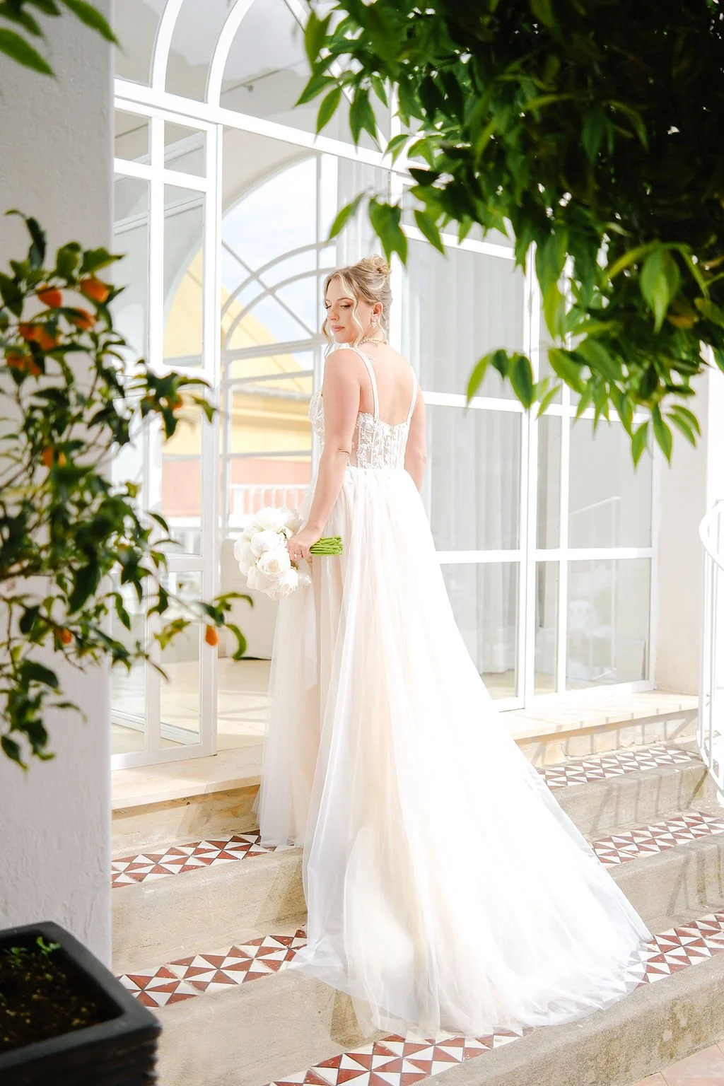 A bride in a white wedding dress holding a bouquet, standing on steps near large glass doors with plants around.