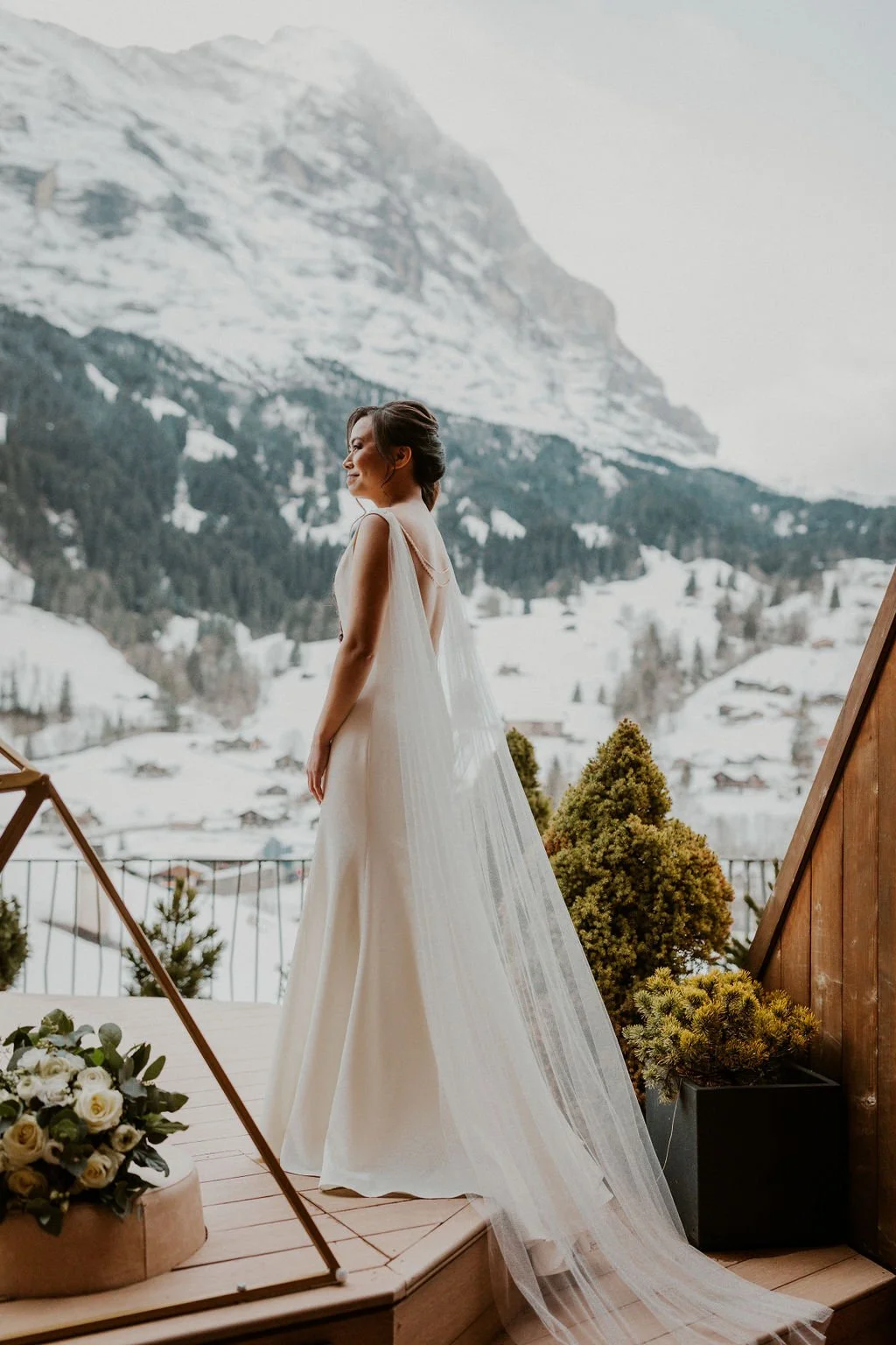 A bride in a white wedding dress with a long veil standing on a wooden balcony overlooking snow-covered mountains and trees.