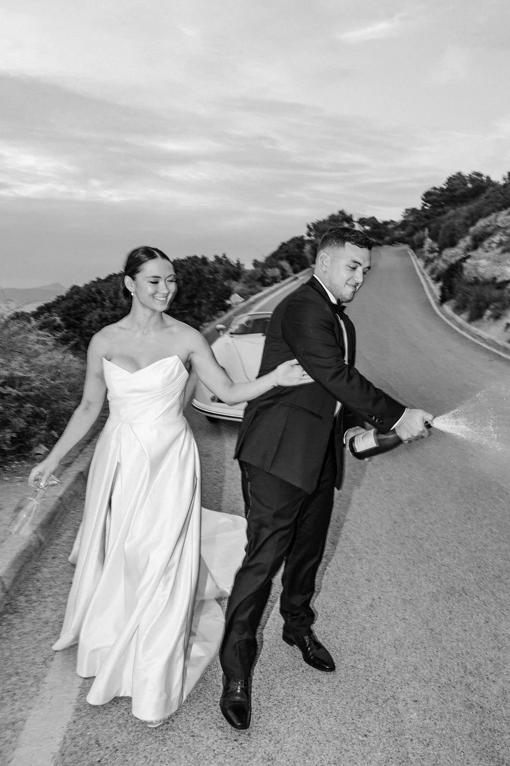 A bride and groom celebrating on the road, with the groom spraying champagne and the bride smiling, set against a scenic outdoor backdrop.