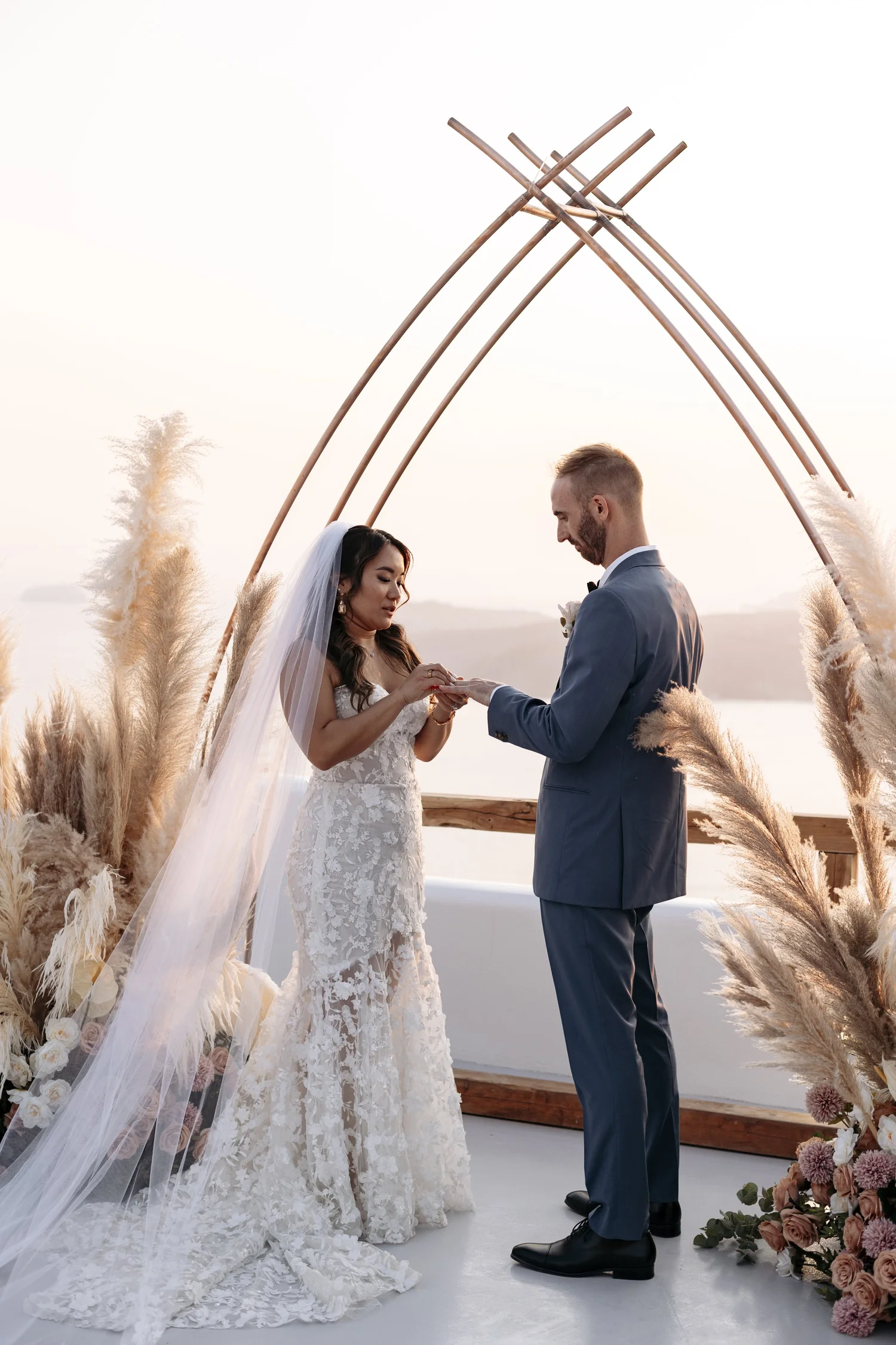 A couple getting married outdoors during sunset, with the bride wearing a lace wedding gown and veil, and the groom in a blue suit, standing under a decorative wooden arch with pampas grass and flowers.