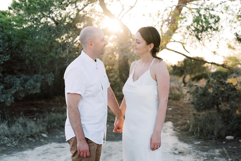 A woman in a white dress and a man in a white shirt hold hands and smile at each other outdoors during sunset, with trees in the background.