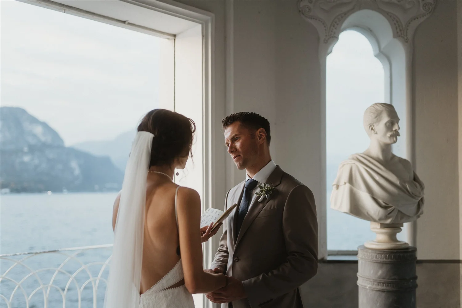 A wedding ceremony taking place indoors with a bride and groom facing each other, holding hands, with a scenic water and mountain view through large windows. There is a white marble bust sculpture on a pedestal to the right.