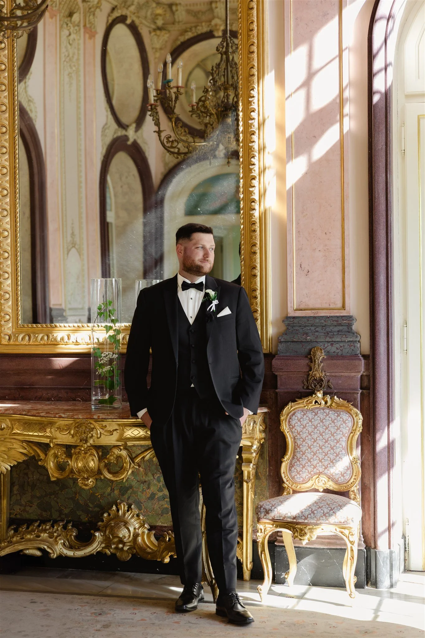 A man in a black tuxedo with a white bow tie and boutonniere stands in an ornate, classical interior with gold-framed mirror and antique furniture, gazing contemplatively to the side.
