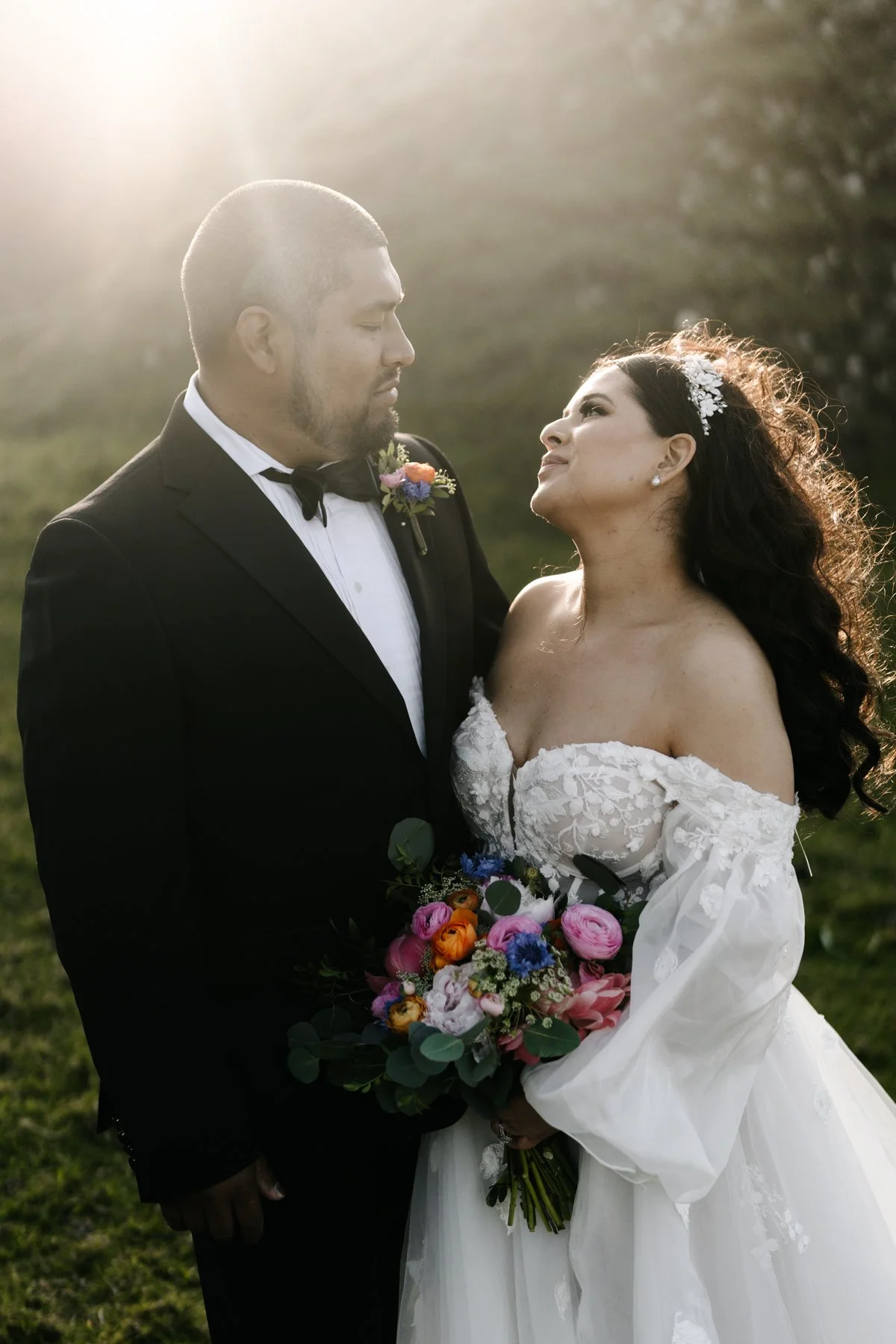 A bride and groom stand close together outdoors at sunset. The bride is holding a colorful bouquet of flowers, and the groom is dressed in a black tuxedo with a bow tie and boutonniere. The bride wears an off-the-shoulder white wedding gown with lace