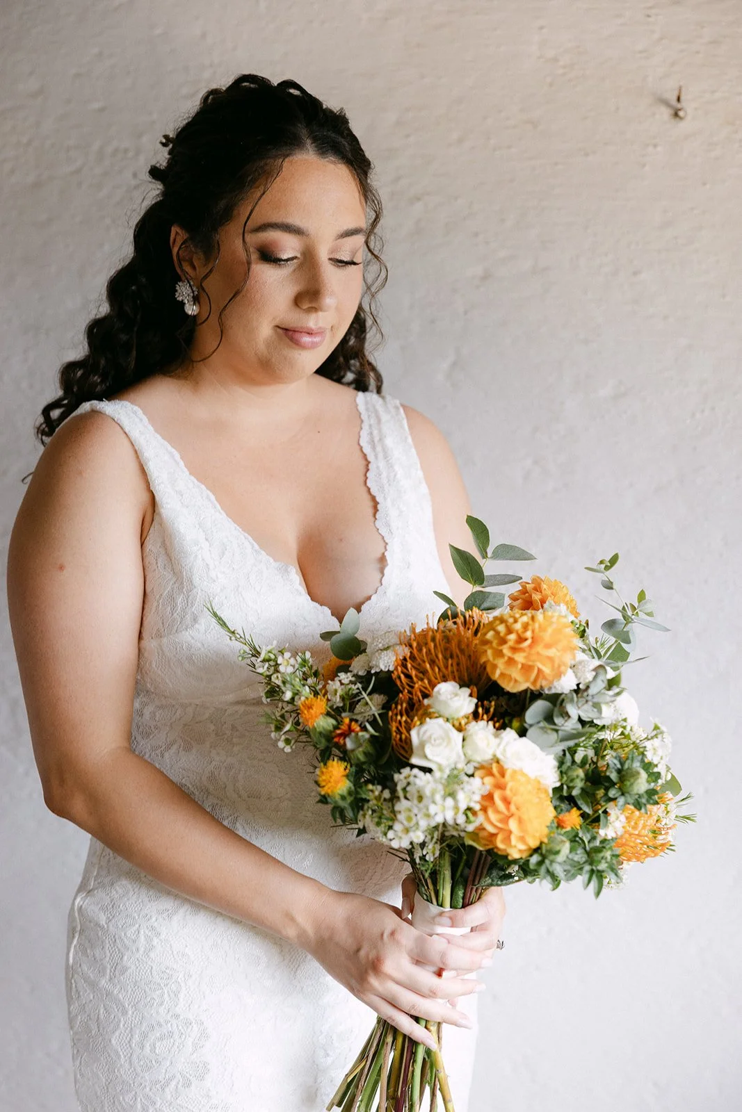 A woman in a white lace dress holding a bouquet of orange, white, and green flowers with closed eyes and relaxed expression.