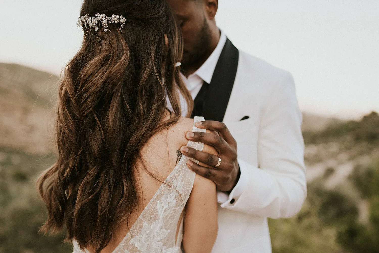 A couple dressed in wedding attire sharing a kiss outdoors. The woman has long wavy brown hair with a decorative hairpiece and wears a lace wedding dress. The man is wearing a white suit with a black lapel.