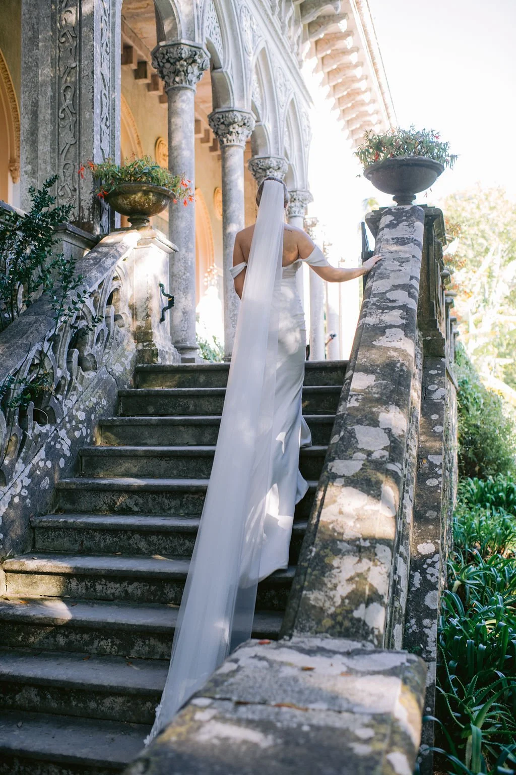 A woman in a white wedding dress ascending stone steps outside an ornate building, with decorative pillars and large planters on either side.