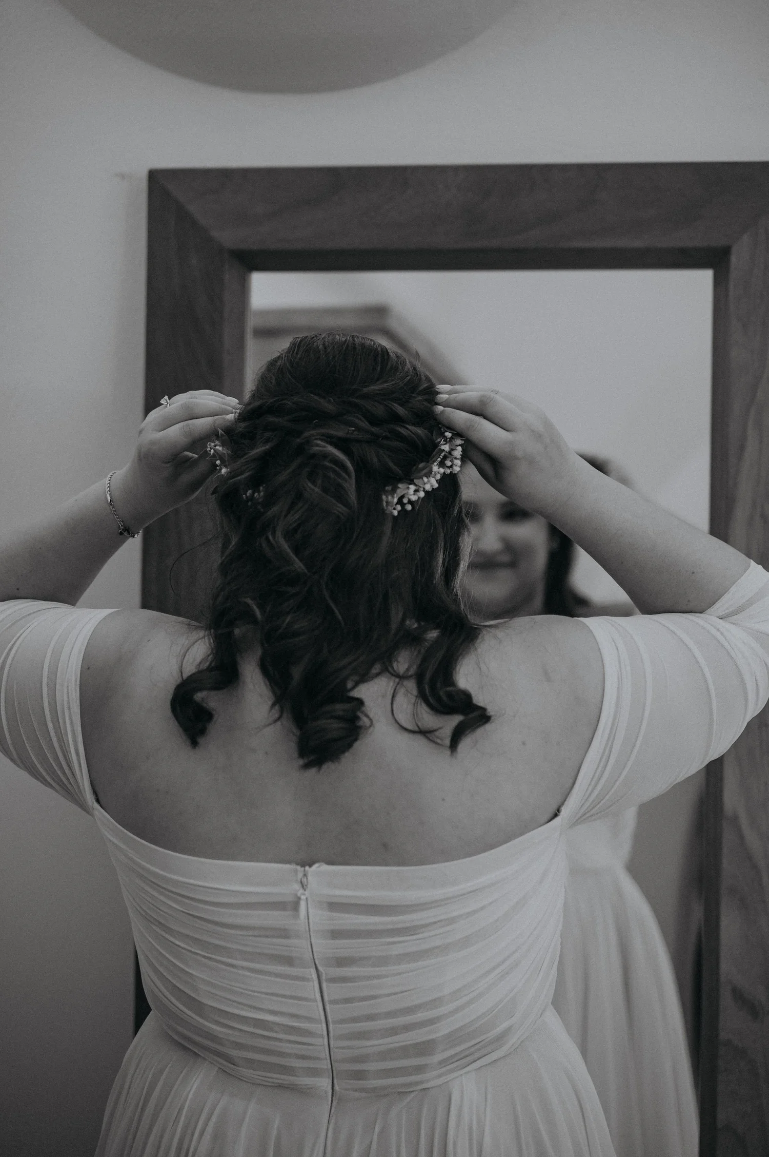 A woman wearing a white dress is seen from the back, adjusting her hair with both hands. She has long, curly hair, and a decorative hairpiece. She is standing in front of a mirror with a dark wooden frame, and the background is blurred.