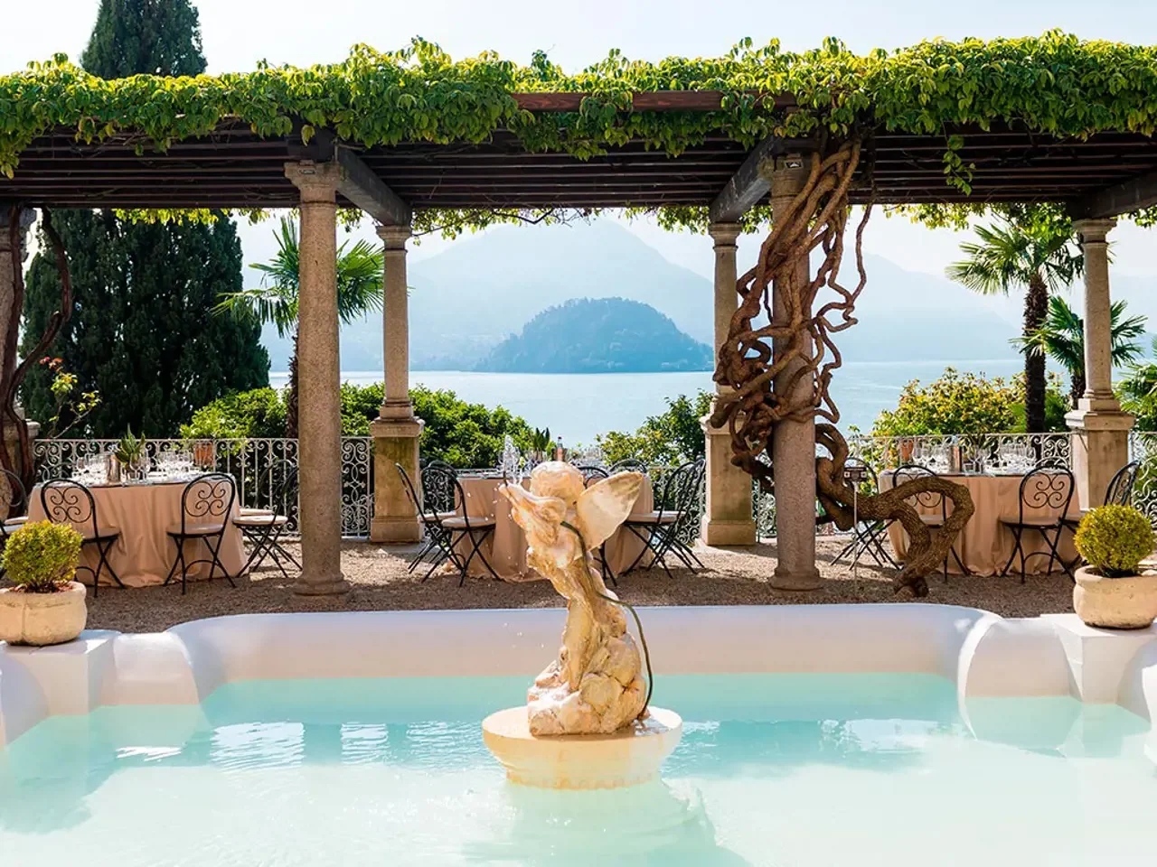 Outdoor dining area overlooking a lake or sea with mountain in the background, featuring a waterfall fountain in the foreground, vine-covered pergola, and tables set for a meal.