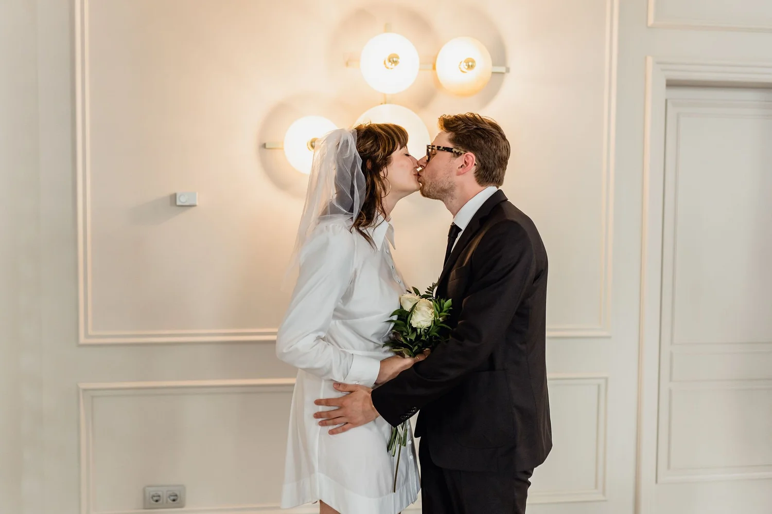 A bride and groom kissing during their wedding, with the bride holding a bouquet of white roses, inside a room with white walls and decorative molding, illuminated by three round wall lamps.
