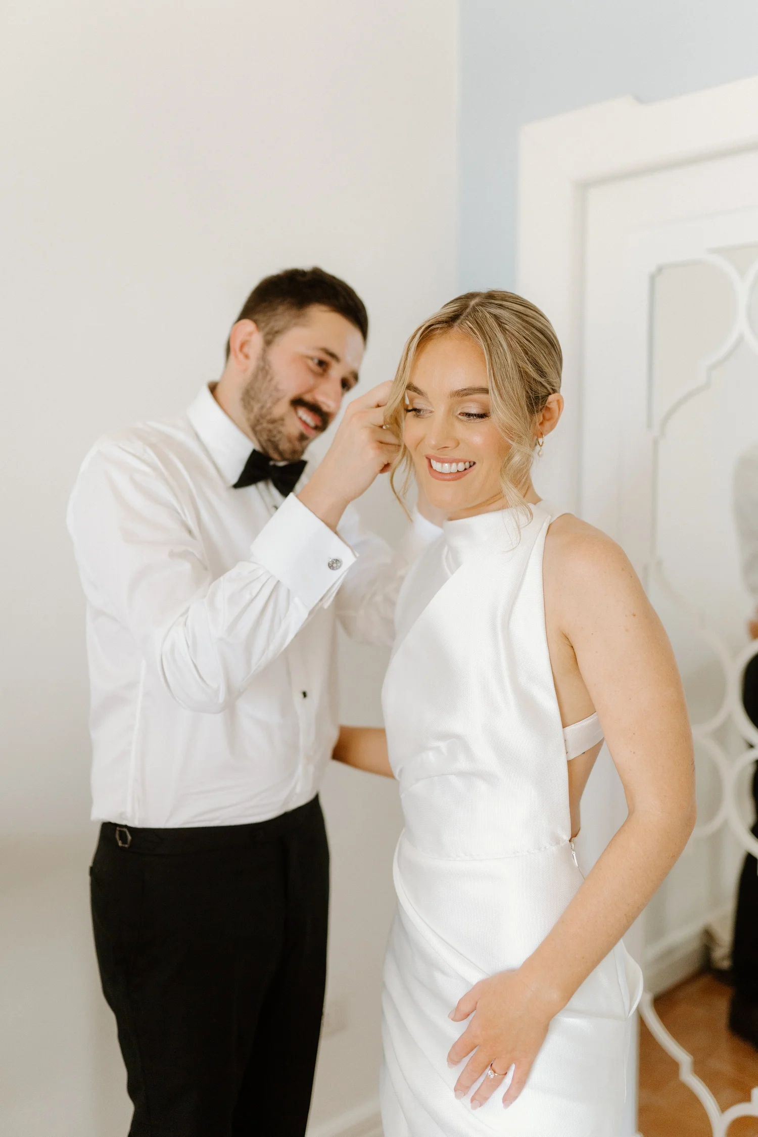 A man in black tuxedo and woman in white wedding dress smiling as he adjusts her hair in a bright, white room.