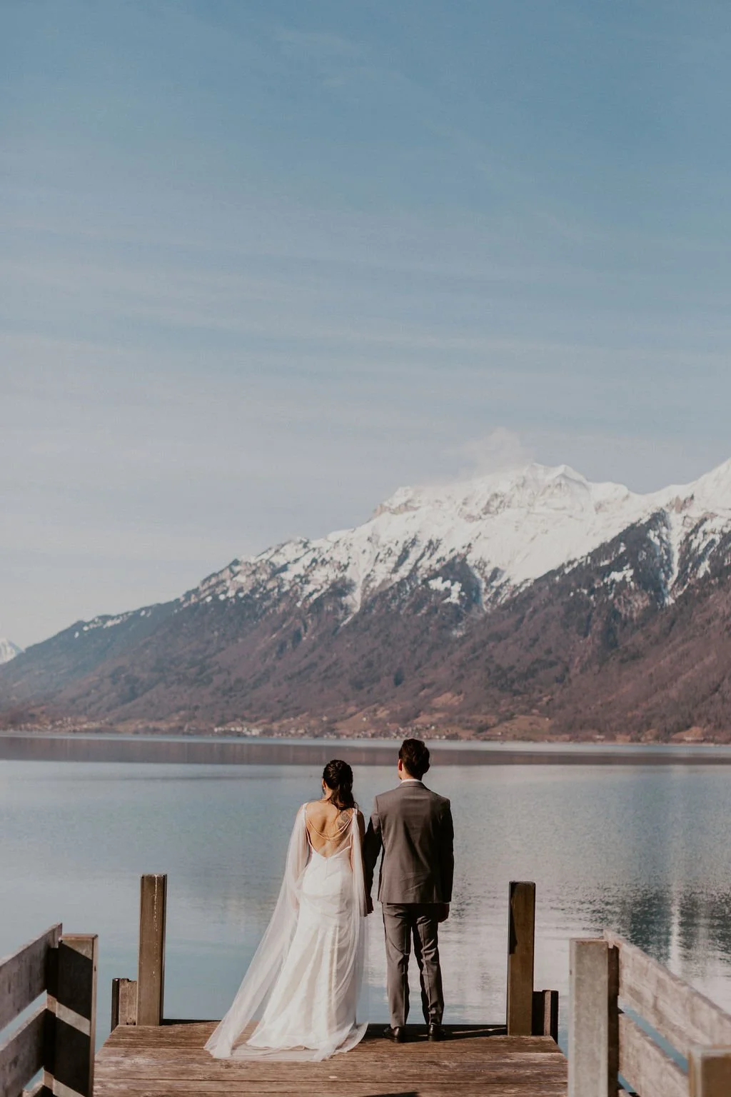 A bride in a white wedding dress and a groom in a gray suit hold hands on a wooden dock by a calm lake, with snow-capped mountains in the background.
