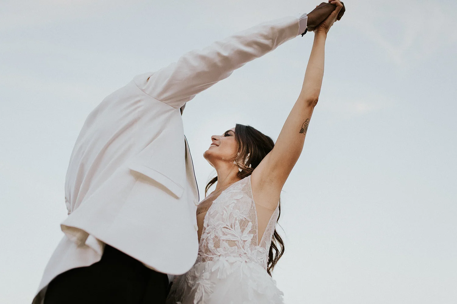 A couple dressed in wedding attire embracing outdoors, with the woman smiling and raising her arm, against a cloudy sky.
