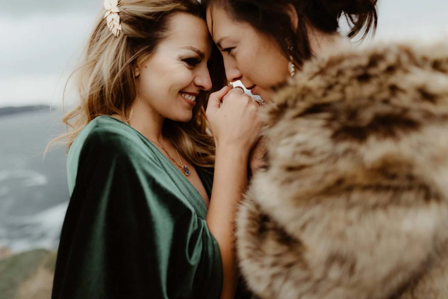 Two women touching foreheads and smiling, with one woman wearing a green velvet top and the other a leopard print coat, outdoors near a body of water.