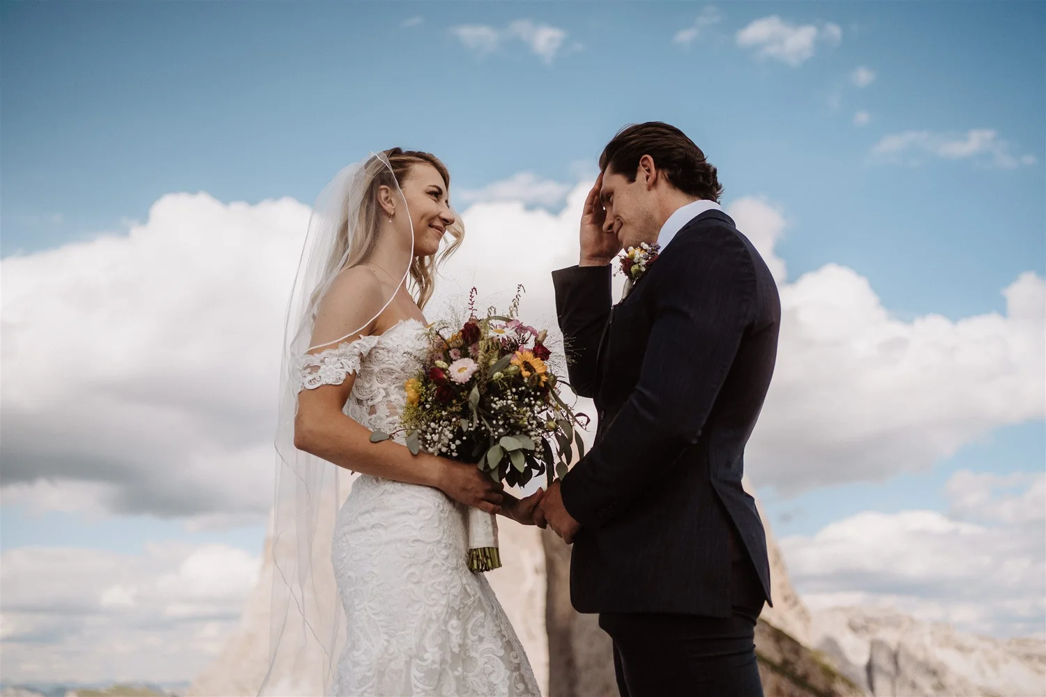 A bride and groom standing outdoors on a cloudy day, exchanging a kiss during their wedding, with the bride holding a colorful bouquet of flowers.