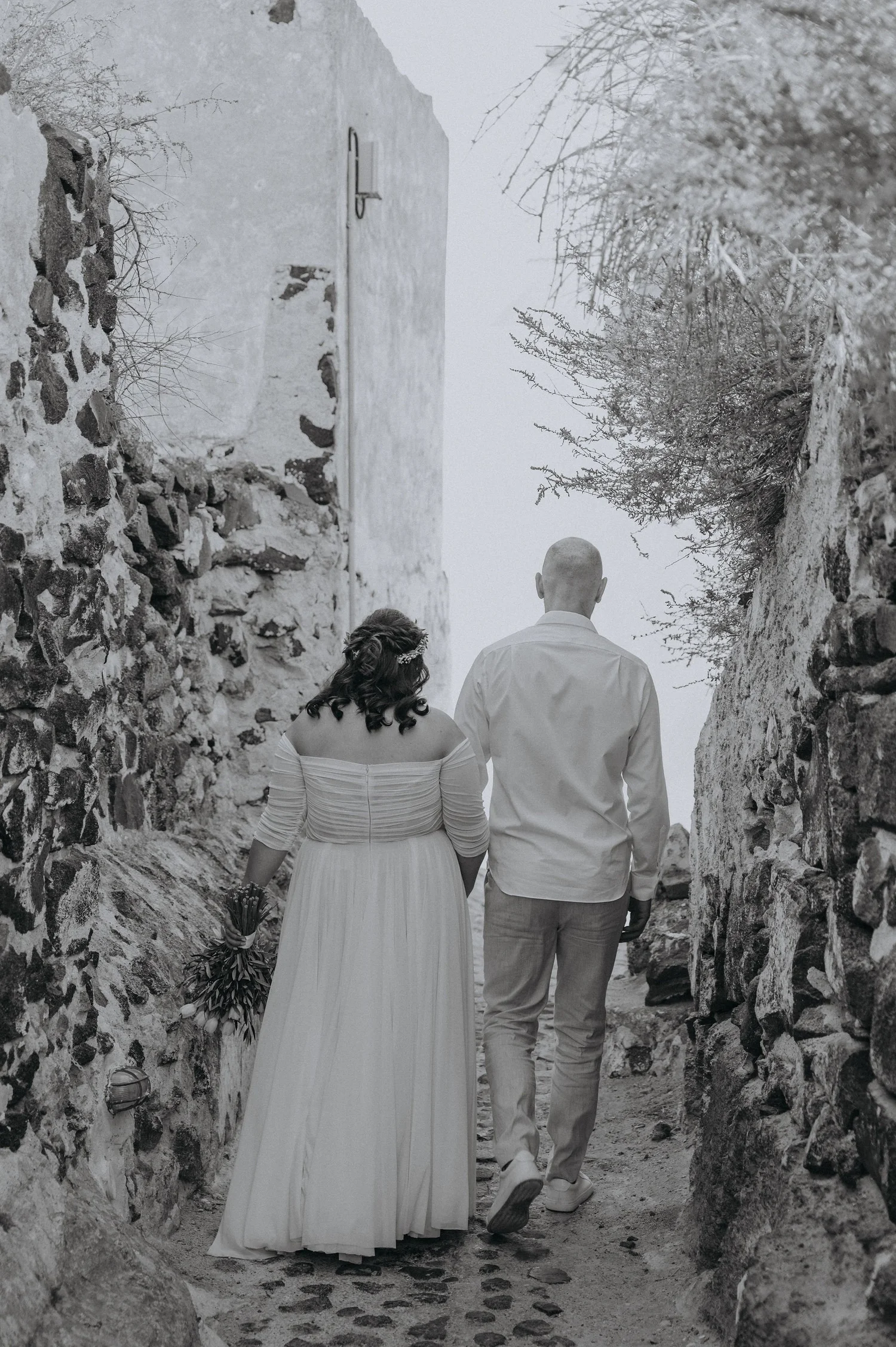 A bride and groom walking hand in hand through a narrow rocky pathway, with the bride holding a bouquet of flowers in her left hand and the groom wearing a white shirt and light-colored pants.