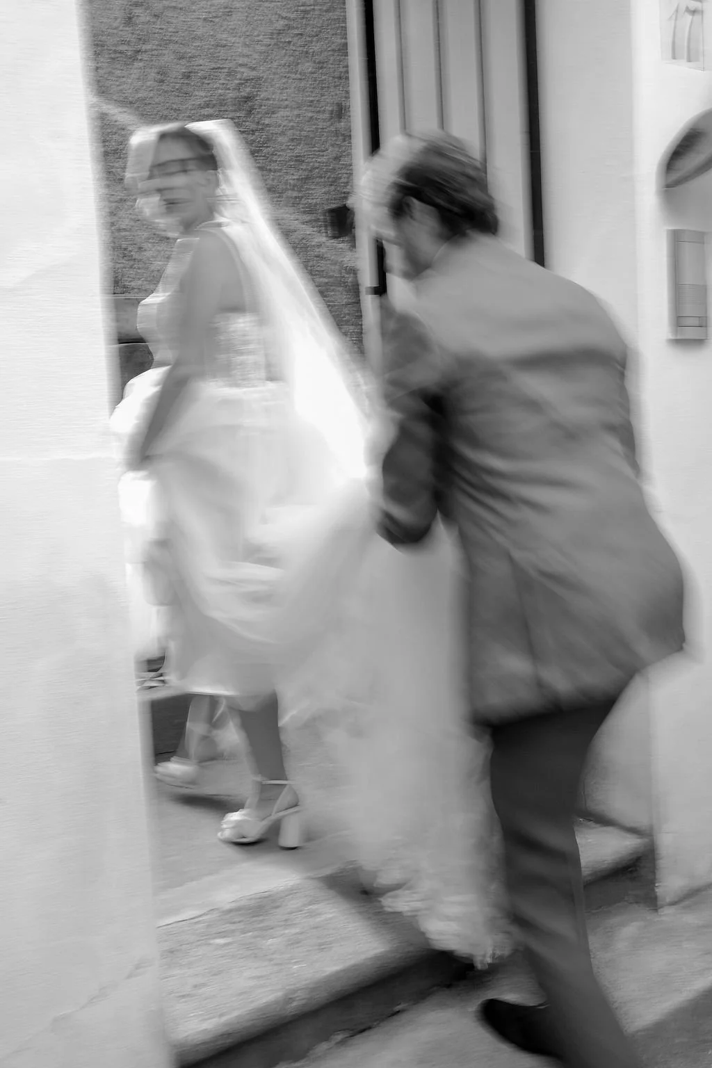 A bride in a wedding dress and veil is seen through a door mirror, with a woman in a coat helping her.