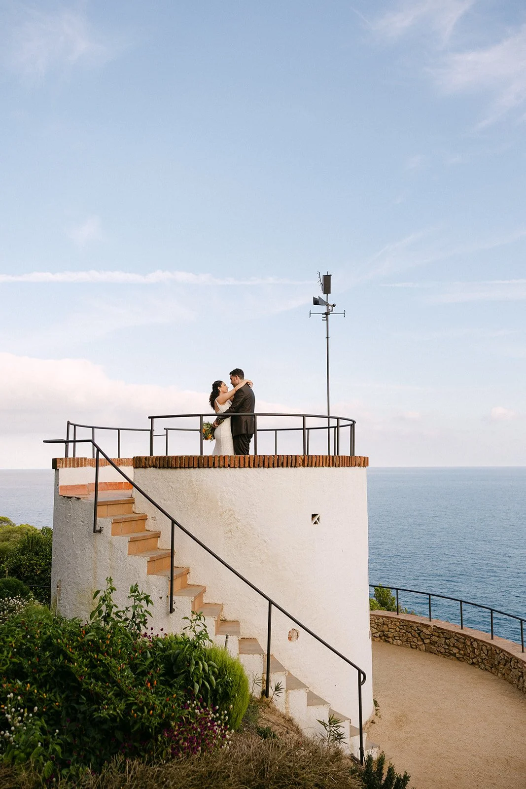 A couple dressed in wedding attire embracing on a rooftop overlooking the ocean, with a blue sky and clouds above, and plants and stairs in the foreground.