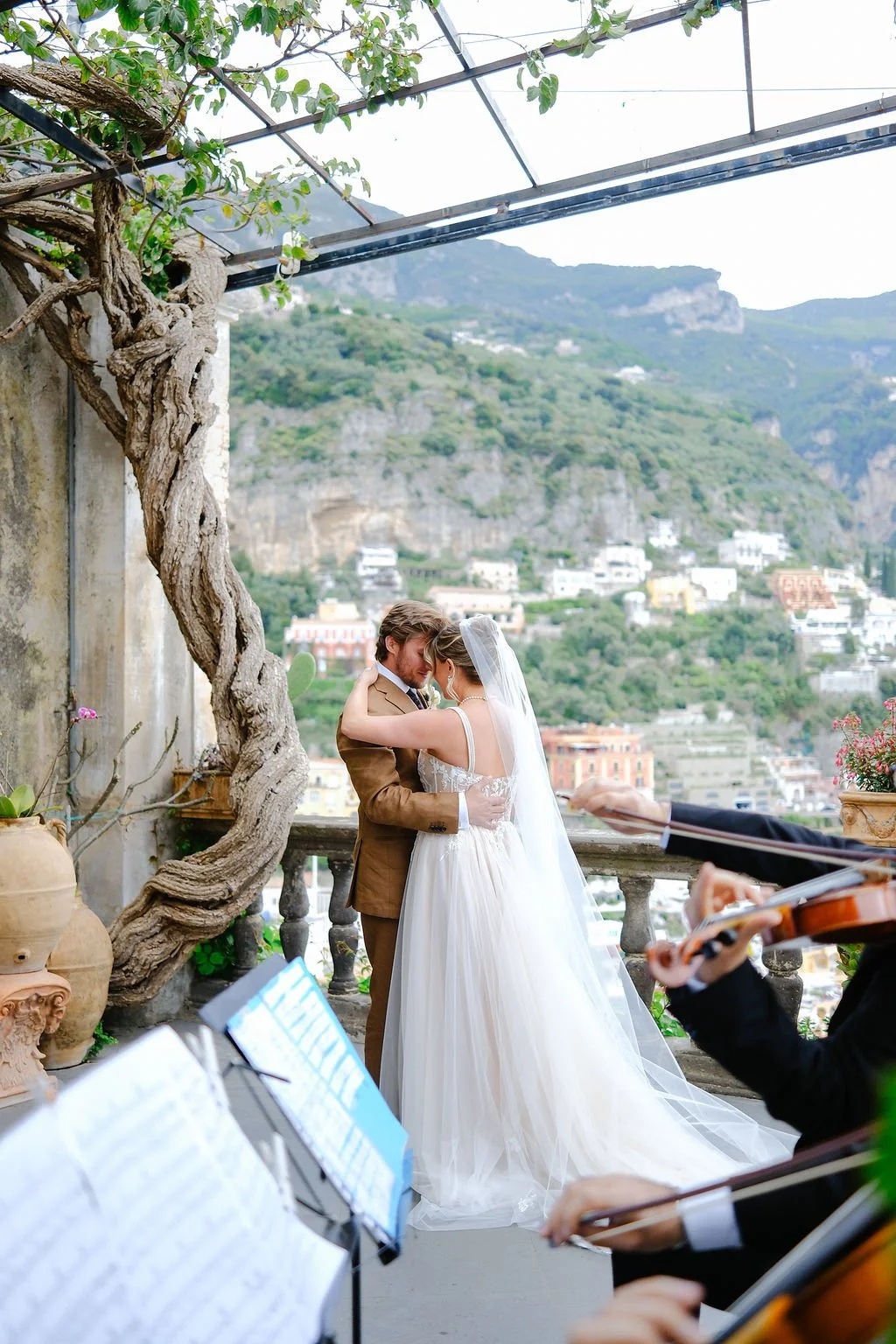 A bride and groom embrace during their wedding ceremony with a scenic hillside landscape in the background, a violinist playing, and wedding music sheets in the foreground.
