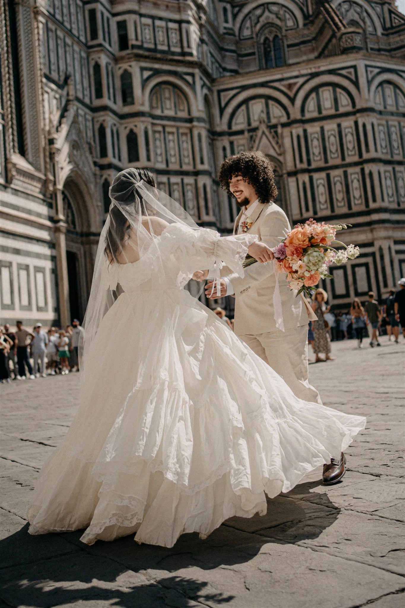 A bride and groom holding hands, dancing in front of a historic cathedral with intricate marble facade, outside on a sunny day. The bride wears a voluminous white wedding dress with a veil, and the groom is in a cream-colored suit holding a bouquet of flowers.