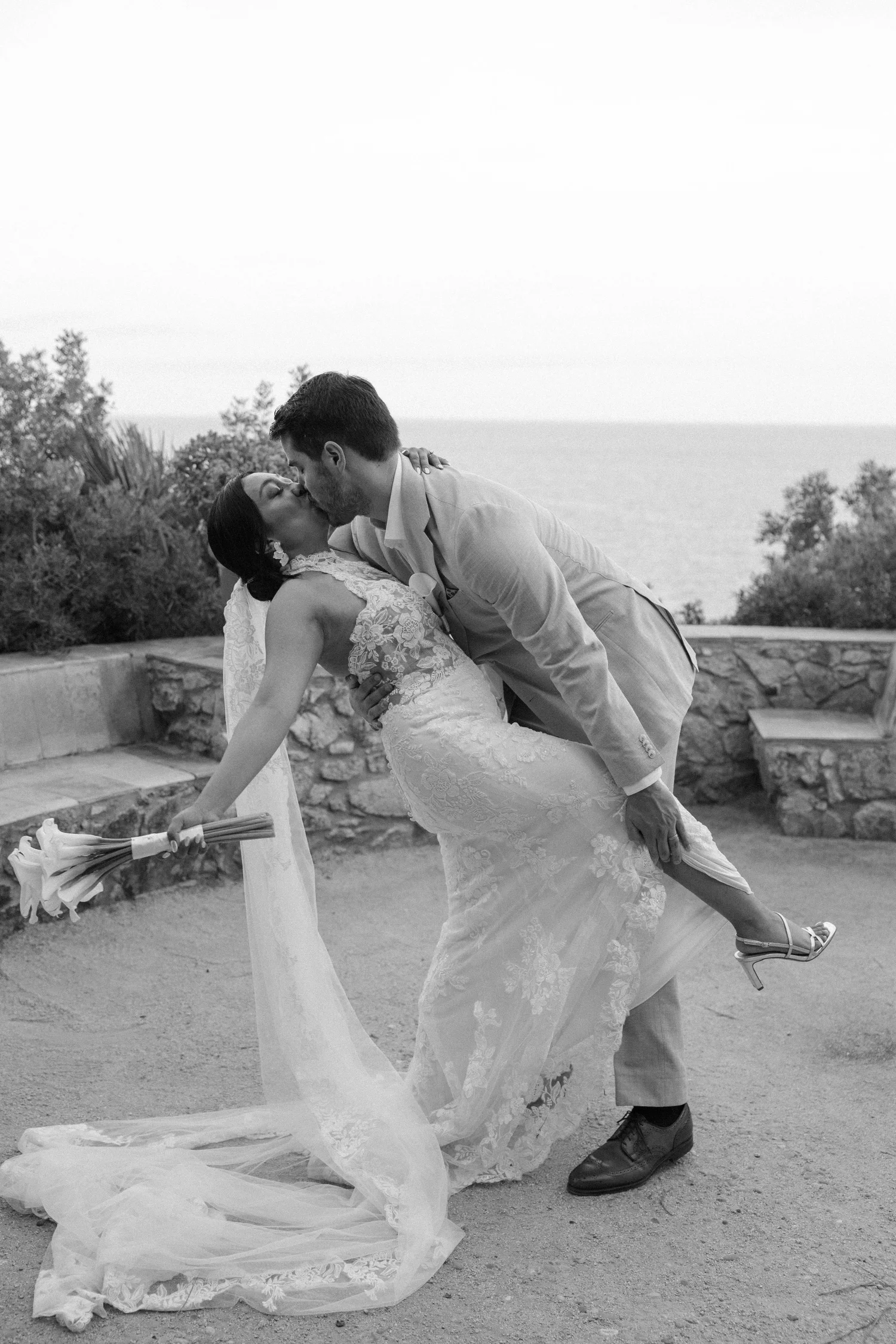 A newlywed couple sharing a romantic moment outdoors, with the groom dipping the bride as they kiss. The bride is holding a bouquet of flowers, and the scene is set against a background of trees and the ocean.