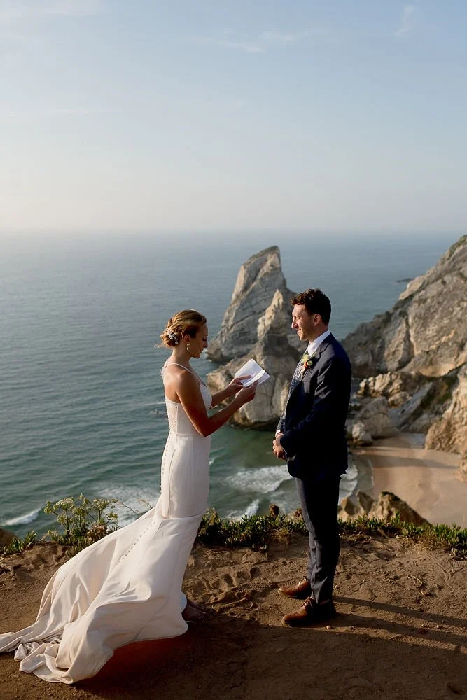 A couple getting married on a cliff overlooking the ocean, with a rocky shoreline and blue sky in the background. The bride wears a white wedding dress, and the groom is in a dark suit. The bride is reading vows from a piece of paper, and the groom i