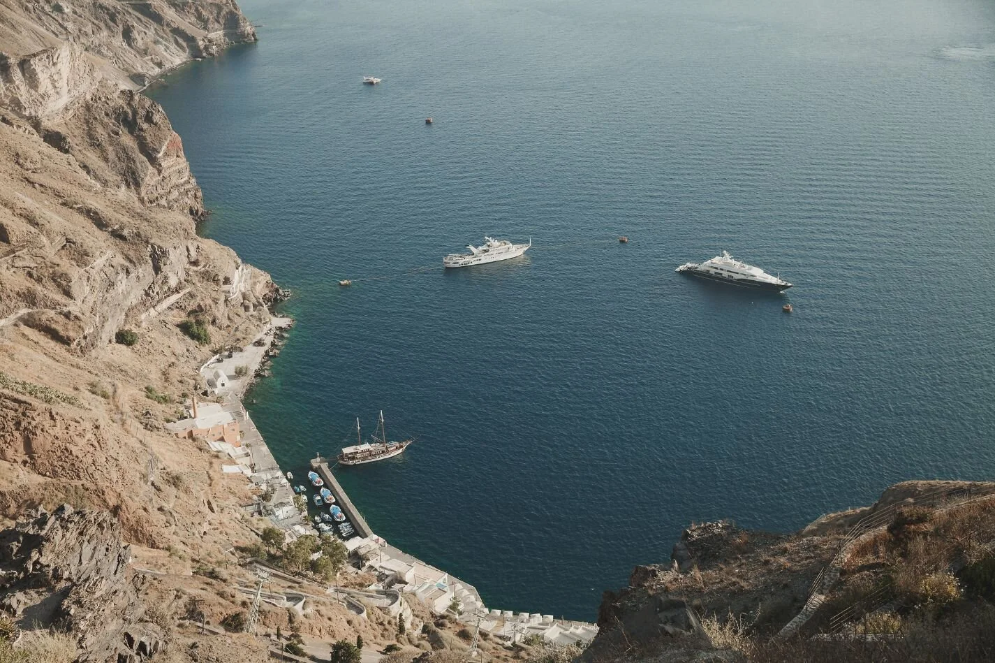 View of boats anchored near a rocky, coastal area with stairs leading down to the water and white buildings nearby.