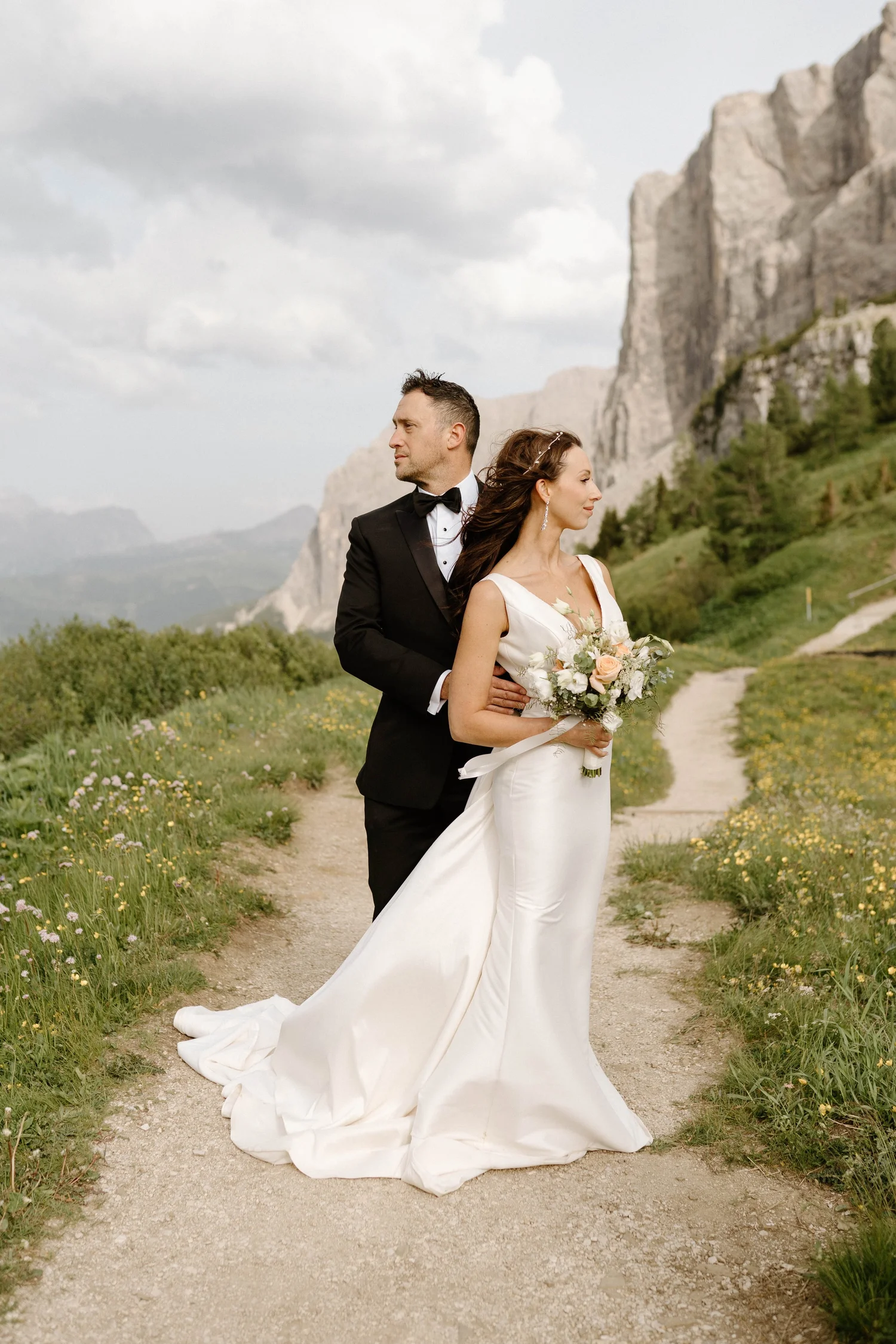A bride and groom standing on a nature trail with green hills and rocky cliffs in the background. The bride is wearing a white lace wedding gown and holding a bouquet of flowers, while the groom is dressed in a black tuxedo with a bow tie.