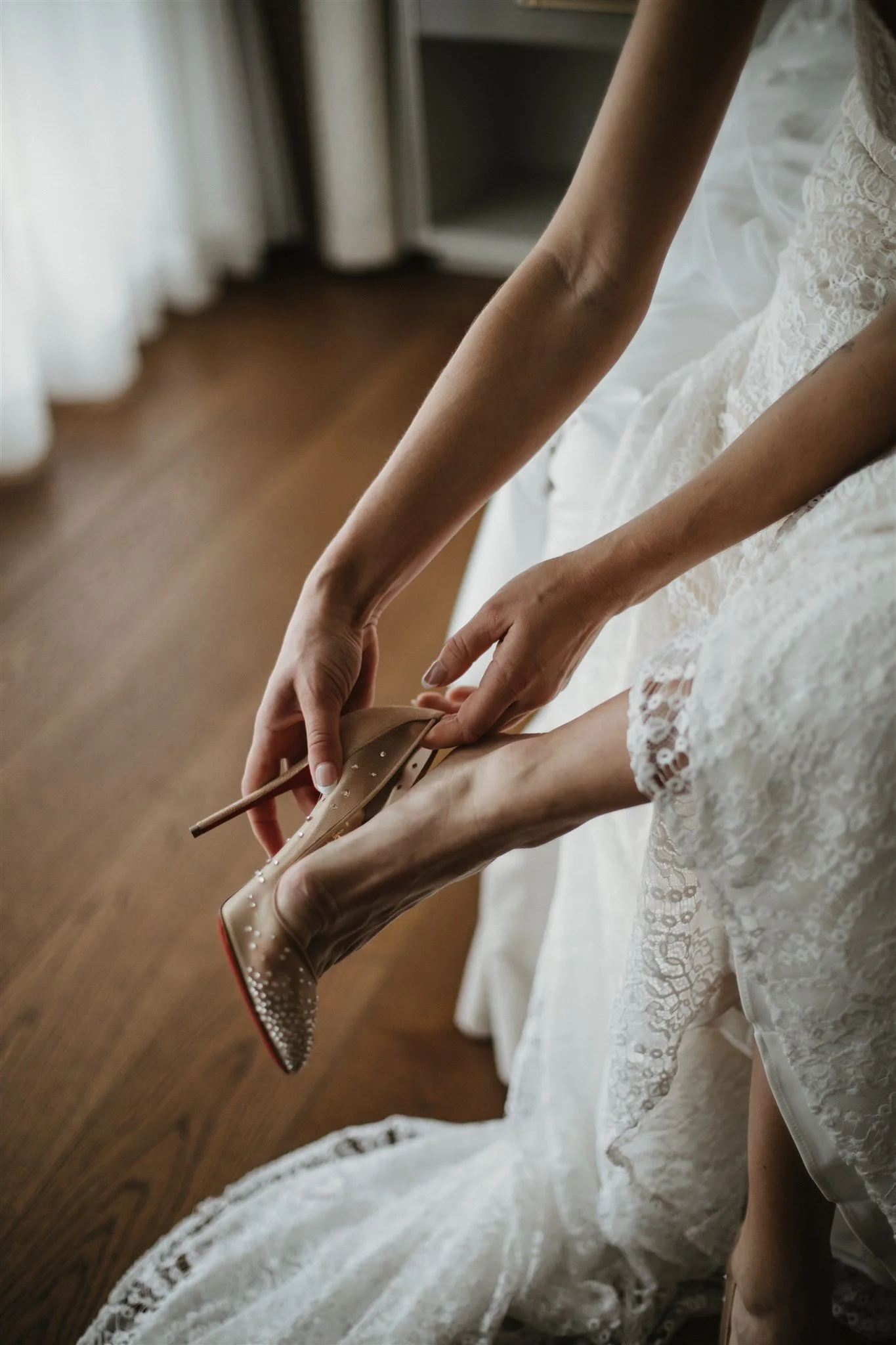 A woman wearing a lace wedding dress is sitting on a wooden floor, adjusting her nude high-heeled shoe.