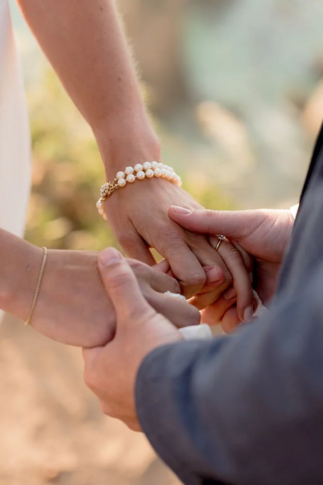 Close-up of couple holding hands during a wedding ceremony, with the bride wearing pearl jewelry and a wedding ring, and the groom wearing a suit and wedding ring, outdoors with blurred natural background.