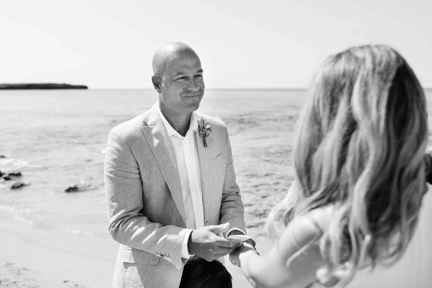 A man in a light-colored suit and white shirt standing on a beach, smiling, during a wedding ceremony, exchanging rings with a woman with long hair.