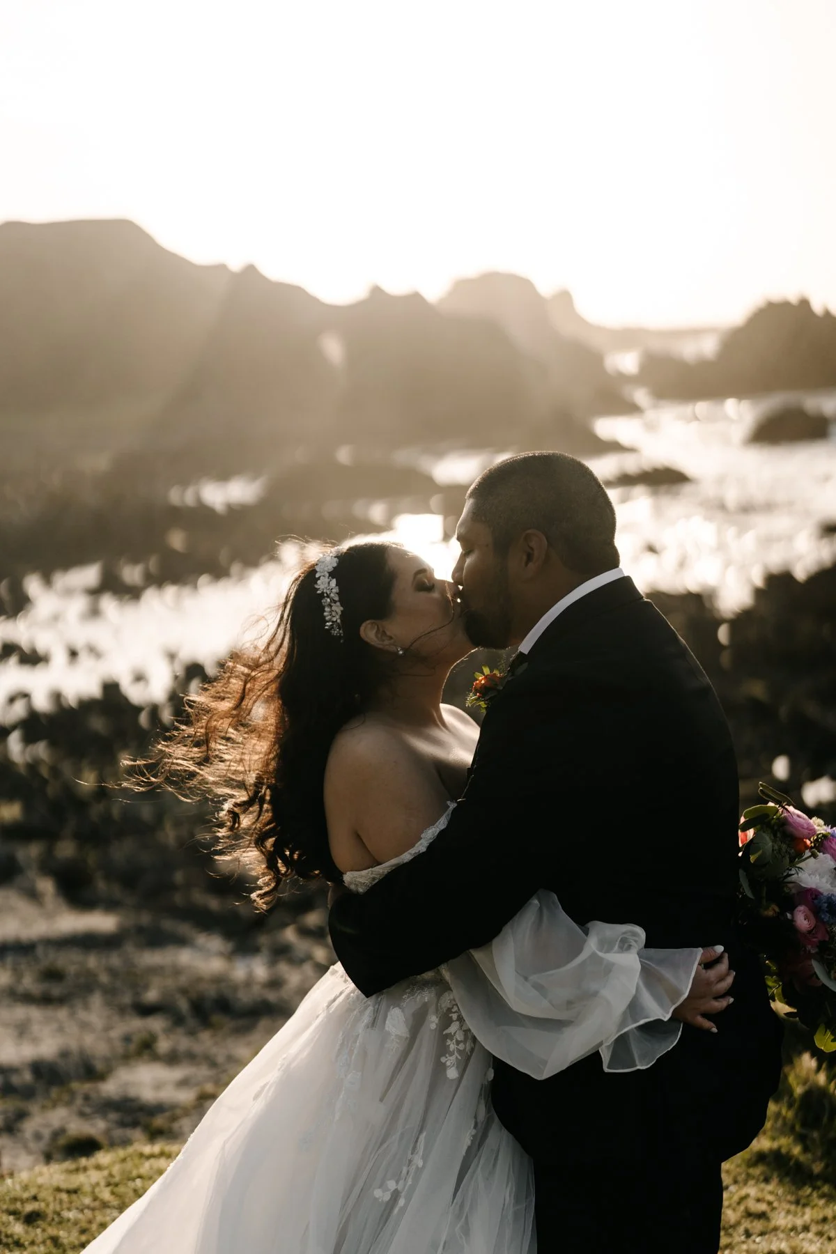 A bride and groom share a kiss outdoors during sunset, with a rocky landscape and water in the background.