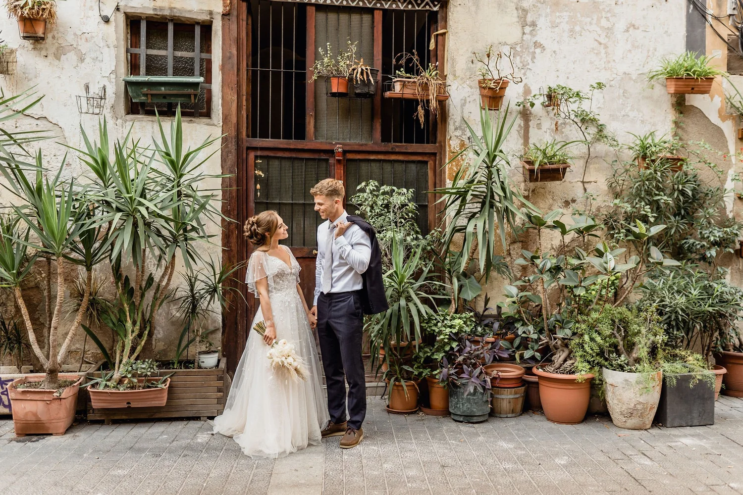 A bride and groom stand facing each other, holding hands, in a rustic outdoor setting surrounded by potted plants and greenery. The bride wears a white wedding dress and holds a bouquet, while the groom is dressed in a white shirt, dark pants, and has a jacket draped over his shoulder. They are smiling at each other against a weathered wall with windows and hanging plants.