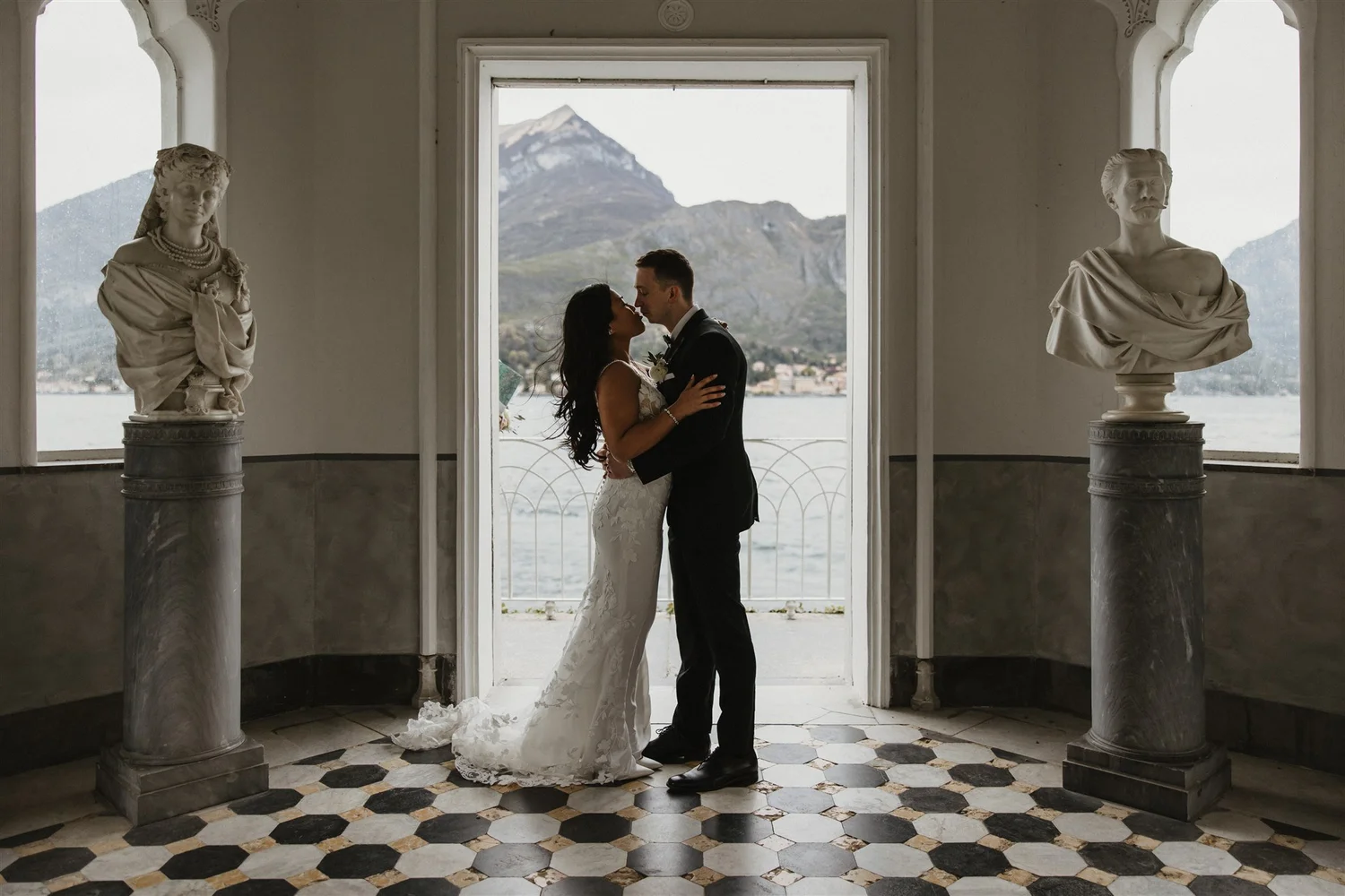 A bride and groom standing close together and about to kiss, inside a building with large windows, overlooking a mountain and water, with two classical statues on pedestals flanking them.