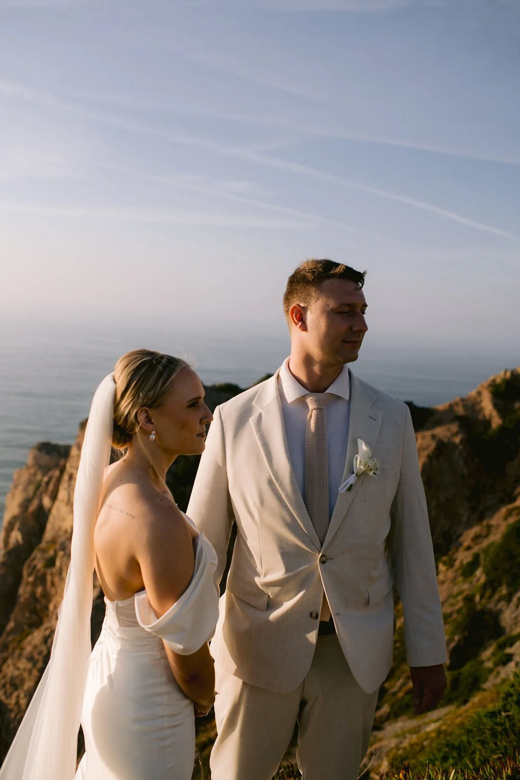 A bride and groom standing outdoors on a hilltop with ocean views. The bride is wearing a strapless wedding dress and veil, and the groom is in a light-colored suit with a tie and boutonniere. It's sunset or late afternoon.
