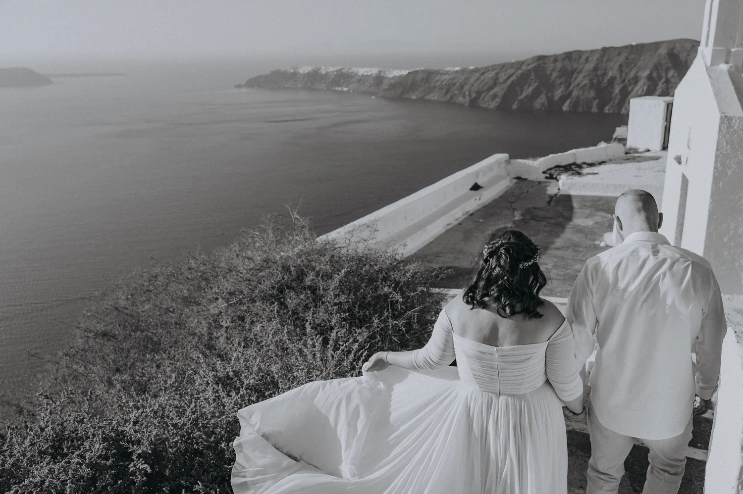 A black-and-white photograph of a couple sitting on a ledge overlooking the ocean and cliffs, with a white building in the foreground.