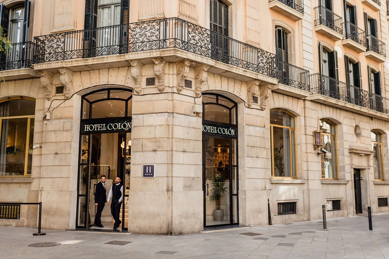 The image shows the entrance of a hotel named "Hotel Colón" on a city street. The building has a classic architectural style with ornate columns and black decorative wrought-iron balconies. Two men are standing at the entrance, dressed in dark suits,