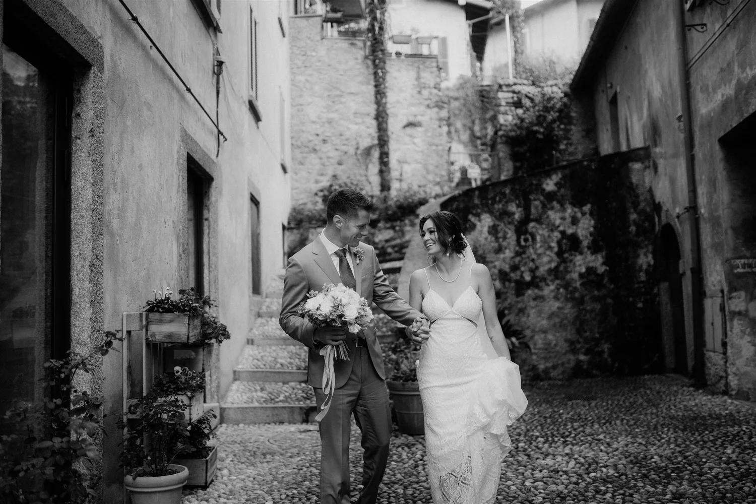 Black and white photo of a wedding couple walking hand in hand down a cobblestone street, smiling at each other. The groom is dressed in a suit and tie, holding a bouquet of flowers, and the bride is wearing a lace wedding gown with spaghetti straps,