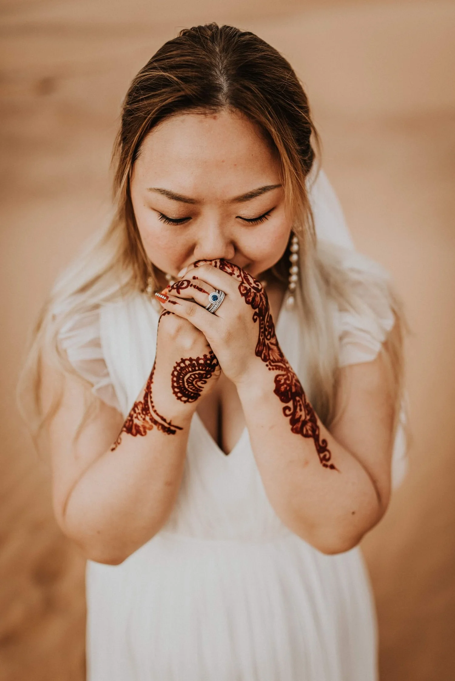 A woman with henna tattoos on her hands and forearms, wearing a white dress, holding her hands together near her face with her eyes closed.