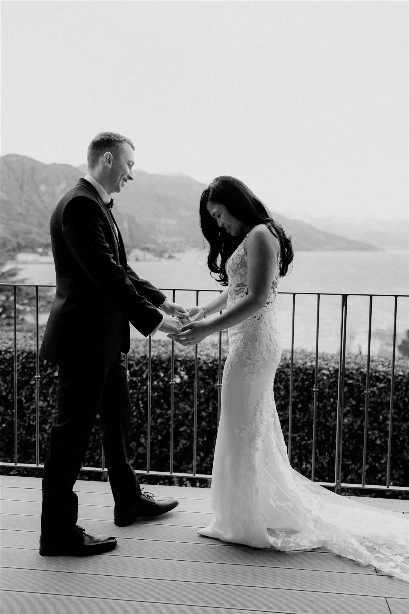 A black-and-white photo of a couple during their wedding ceremony on a balcony with a scenic view of mountains and a body of water in the background. The groom is wearing a tuxedo, and the bride is wearing a lace wedding gown. They are holding hands