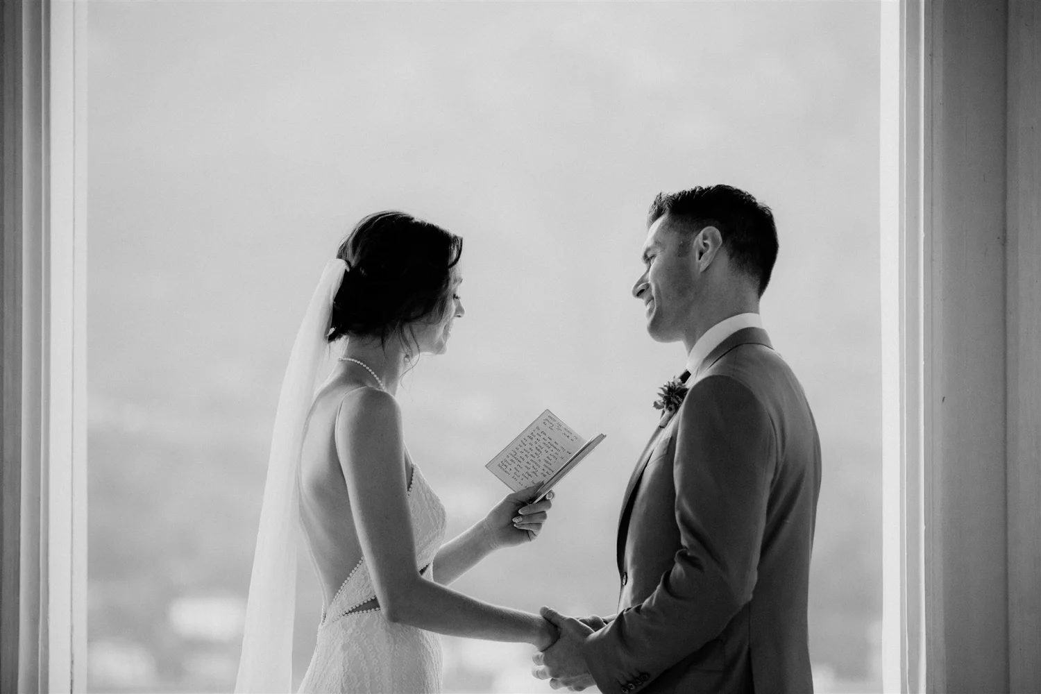 A black and white photo of a bride and groom standing face to face, smiling and holding hands, with the bride reading vows from a small booklet.
